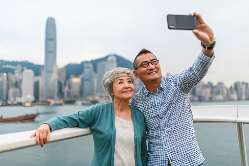 Senior Chinese woman and mature man taking selfie at Ocean Terminal Deck with Hong Kong cityscape in background.
