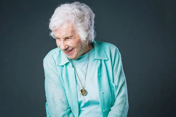 Photo of Close-Up Shot of Sweet, Joyful, Happy, Wise Elderly Senior Caucasian Woman Smiling, Wearing Her Favorite Teal Blue Shirt and Standing Against a Gray Background Looking Down and Admiring Something in Studio with Copy Space
