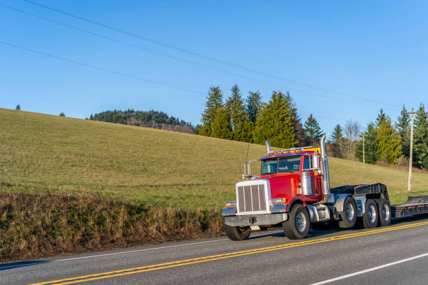 istock Red day cab big rig semi truck with step down semi trailer for carry oversized loads running on the road with field hill on the side 1210006101