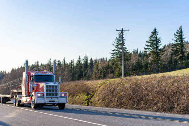 istock Big rig red classic semi truck with large grille guard transporting empty step down semi trailer running on the road to loading side 1210006195