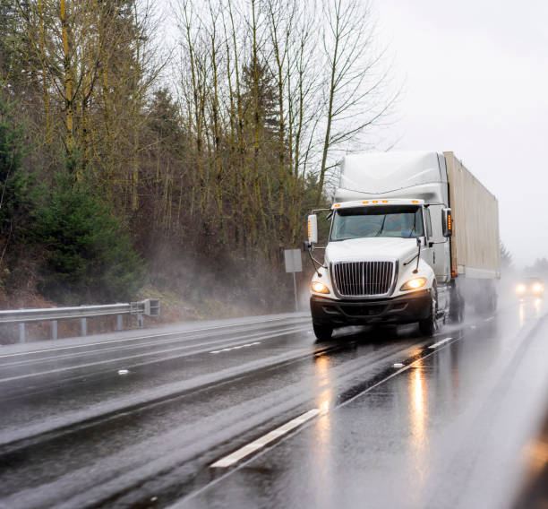 Big rig white diesel semi truck transporting container with commercial cargo running on the wet road at raining weather Big rig white diesel semi truck with turned on headlights transporting container with commercial cargo running on the wet multiline highway at raining weather with reflection on the road surface truck rain stock pictures, royalty-free photos & images