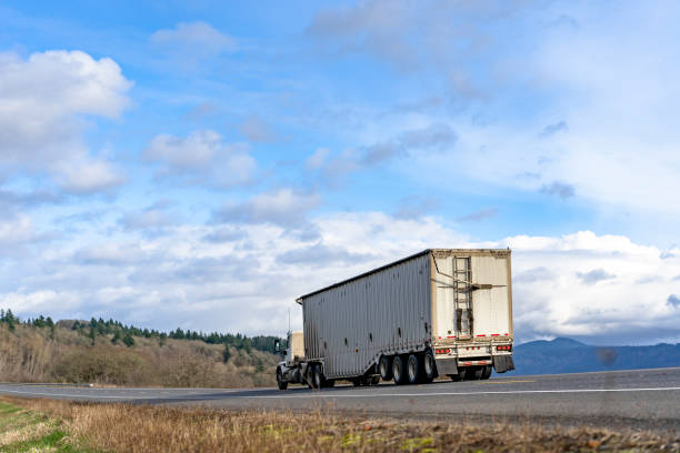 istock Day cab big rig semi truck transporting cargo in covered corrugated bulk semi trailer driving on the road with frees on the hill 1210007267