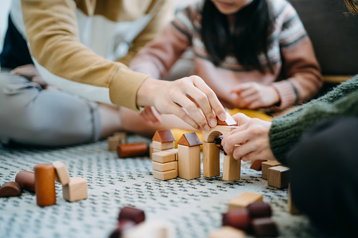 Close up of joyful Asian parents sitting on the floor in the living room having fun and playing wooden building blocks with daughter together