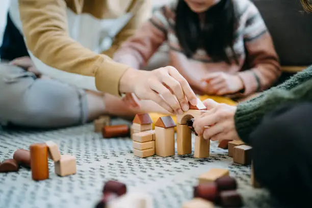 Photo of Close up of joyful Asian parents sitting on the floor in the living room having fun and playing wooden building blocks with daughter together