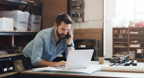 Designing dreams is what he does best Shot of a young businessman using a smartphone while designing a blueprint in a creative office warehouse office stock pictures, royalty-free photos & images