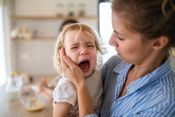 una madre che tiene in braccio una figlia bambina che piange al chiuso in cucina. - piangere foto e immagini stock