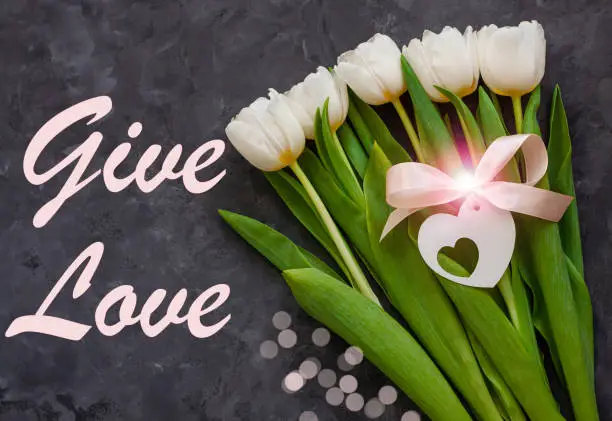 Give Love card message sign. Giving Tuesday Charity Donations concept. White tulip flowers and heart card tag with pink ribbon bow dark background flat lay. Flower Bouquet with bokeh lights top view.