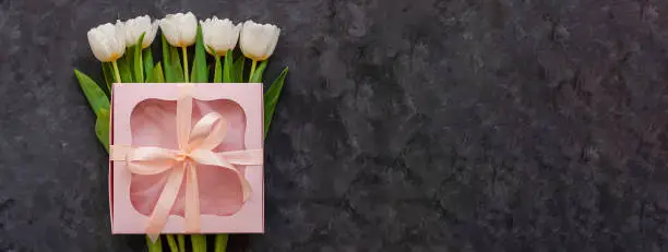White tulip flowers and pink gift box with ribbon bow on dark background flat lay site header. Place for text 8 March Happy Womens Mothers Day.Flower Bouquet greeting card.Copy space website banner