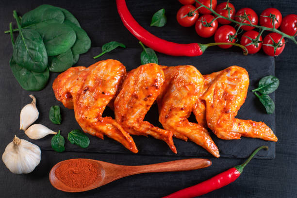 Raw pickled chicken wings on a black plate with red hot pepper and cherry tomatoes on a wooden black background. Chicken marinated meat.