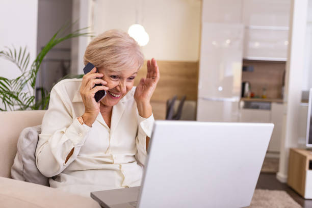 istock Happy senior Woman with Laptop Computer, Sitting at the Couch While Talking to Someone on Phone. Using from new technology is easier and faster 1210043539
