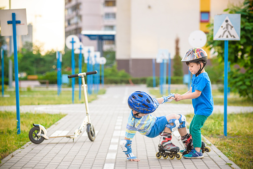 Two boys sibling brothers together in park, helps boy with roller skates to stand up after fall. Friendship and active leisure summer holidays time with family concept.