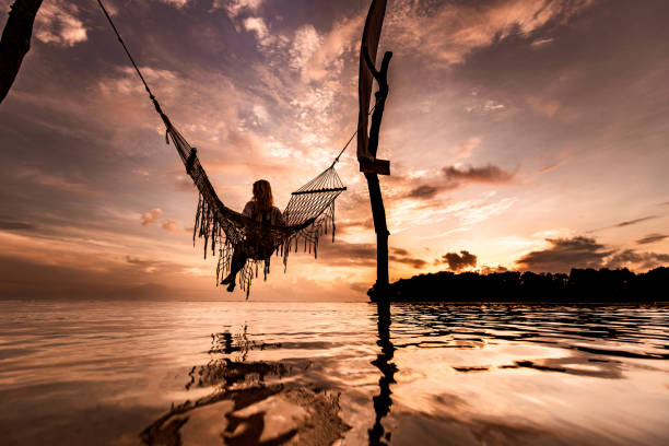 vue arrière d’une femme s’asseyant dans le hamac au-dessus de la mer au coucher du soleil. - hammock beach vacations tropical climate photos et images de collection