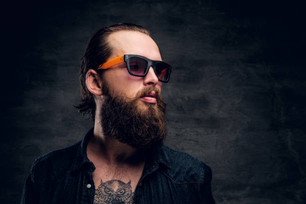 Bearded man in sunglasses is posing for photographer at dark photo studio. Handsome bearded man in sunglasses is posing for photographer at dark photo studio. chest tattoos for men designs stock pictures, royalty-free photos & images