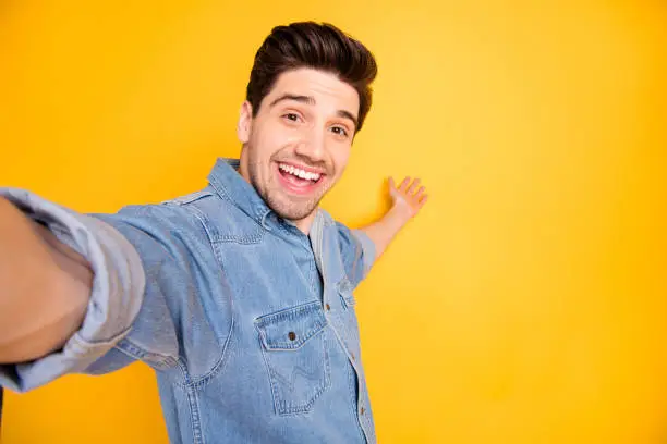 Photo of cheerful positive attractive man welcoming you to come to empty space, behind him taking selfie isolated vivid color background