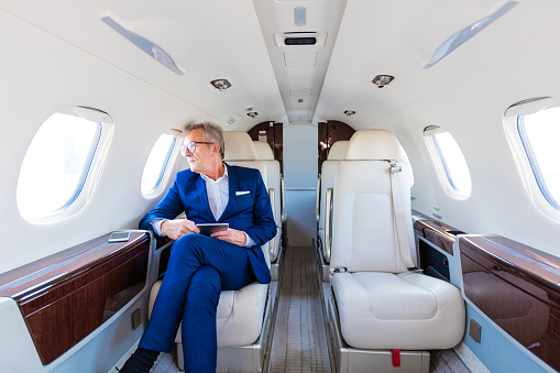 Senior businessmen wearing suit, sitting in private airplane, holding digital tablet, looking out the window.