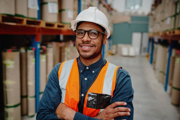 istock Portrait of handsome cheerful supervisor in warehouse standing with folded hands with material while wearing orange vest and white hardhat 1210069333
