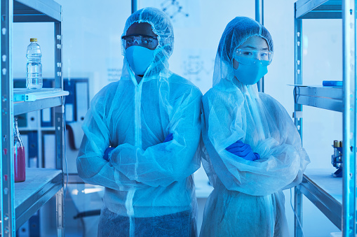 Horizontal medium portrait shot of two unrecognizable laboratory scientists in protective workwear standing with arms crossed looking at camera