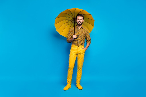 Full body photo of cheerful dreamy energetic guy fun free time on rainy weather, hold colorful bright parasol wear total yellow look sneakers isolated over blue color background