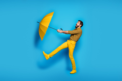Full body profile side photo of scared frustrated anxious man travel windy weather, catch flying bright parasol wear checkered trousers sneakers isolated over blue color background
