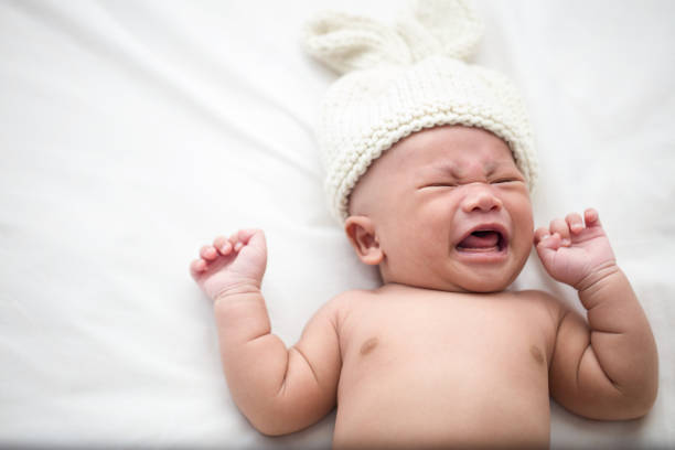 a 1-month-old baby is lying and crying because of feeling hungry on a white mattress. the infant feels unwell due to sleepiness. - baby lying down sleeping asian ethnicity imagens e fotografias de stock