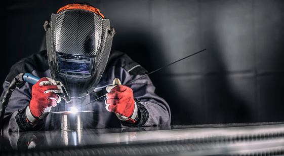 Industrial welder with a torch and protective helmet welding metal profiles in factory