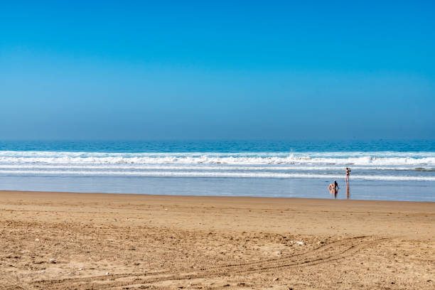 Atlantic Ocean at Taghazout, a tourist area in Morocco that is receiving considerable investment in new hotels, apartments etc. In the midground are 2 European women sitting and standing in the surf.