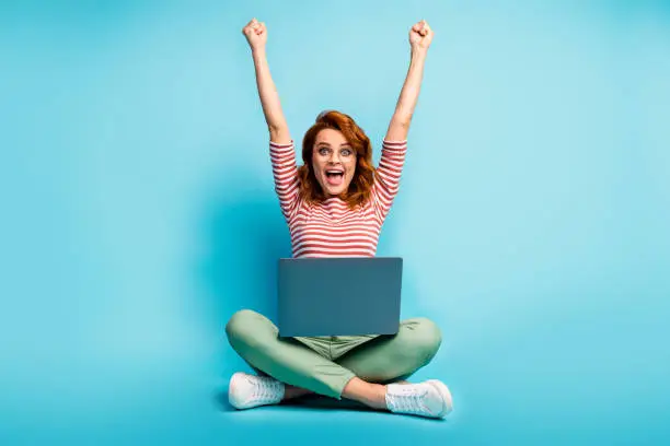 Photo of Full size photo of excited ginger hair woman sit floor legs crossed work laptop finish start-up report scream yeah raise fists wear green white sweater footwear isolated blue color background