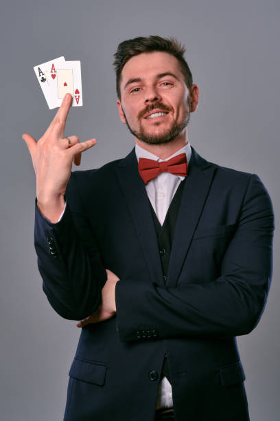 istock Man in black classic suit and red bow-tie showing two playing cards while posing against gray studio background. Gambling, poker, casino. Close-up 1210115944