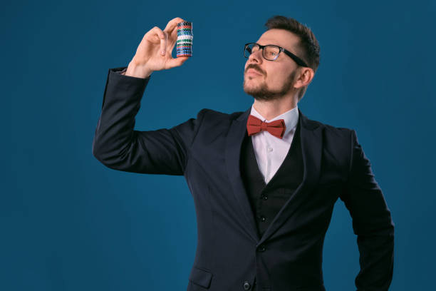 istock Man in black classic suit, red bow-tie, glases is showing some colored chips, posing on blue studio background. Gambling, poker, casino. Close-up 1210116656