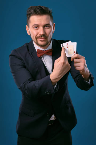 istock Man in black classic suit and red bow-tie showing two playing cards while posing against blue studio background. Gambling, poker, casino. Close-up 1210116887