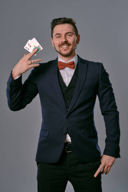 istock Man in black classic suit and red bow-tie showing two playing cards and chips, posing on gray studio background. Gambling, poker, casino. Close-up 1210118373