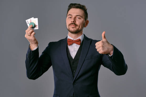 istock Man in black classic suit and red bow-tie showing two playing cards and chips, posing on gray studio background. Gambling, poker, casino. Close-up 1210118389