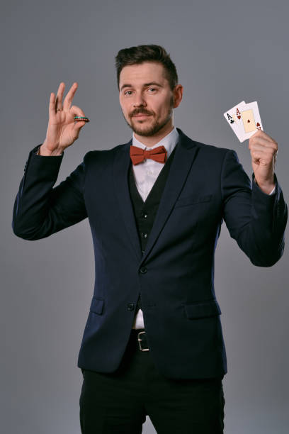 istock Man in black classic suit and red bow-tie showing two playing cards and chips, posing on gray studio background. Gambling, poker, casino. Close-up 1210118448