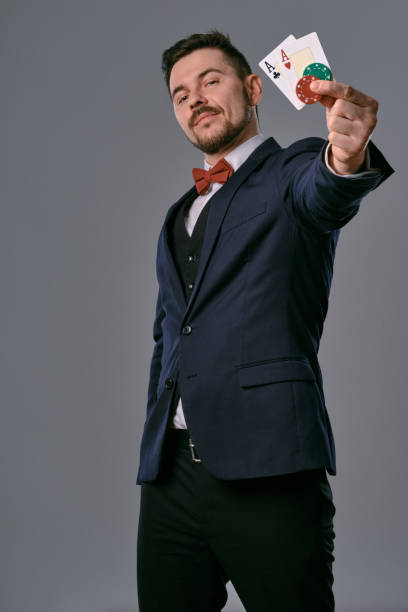 istock Man in black classic suit and red bow-tie showing two playing cards and chips, posing on gray studio background. Gambling, poker, casino. Close-up 1210118483