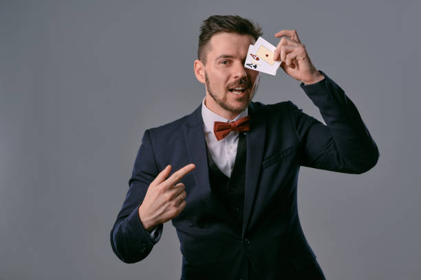 istock Man in black classic suit and red bow-tie showing two playing cards while posing against gray studio background. Gambling, poker, casino. Close-up 1210118851