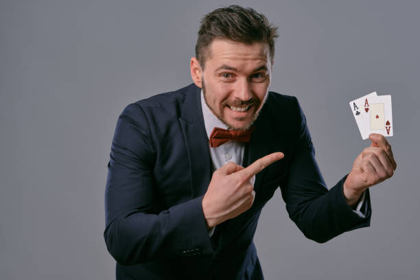 istock Man in black classic suit and red bow-tie showing two playing cards while posing against gray studio background. Gambling, poker, casino. Close-up 1210118859