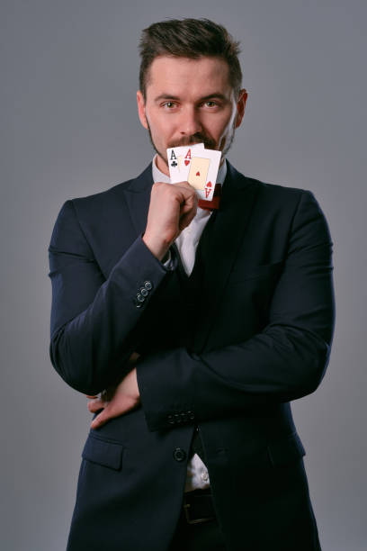 istock Man in black classic suit and red bow-tie showing two playing cards while posing against gray studio background. Gambling, poker, casino. Close-up 1210119794