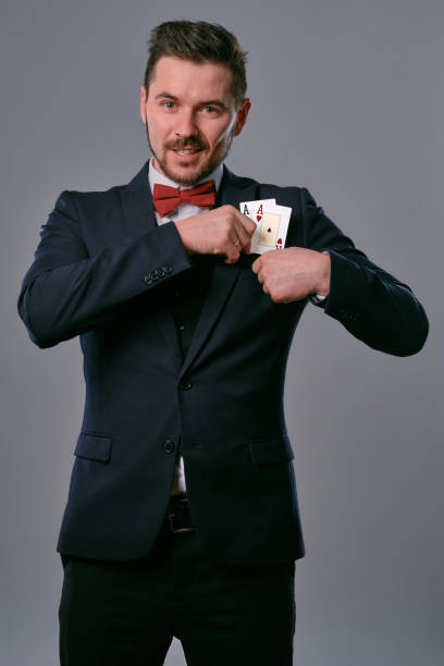 istock Man in black classic suit and red bow-tie showing two playing cards while posing against gray studio background. Gambling, poker, casino. Close-up 1210119803