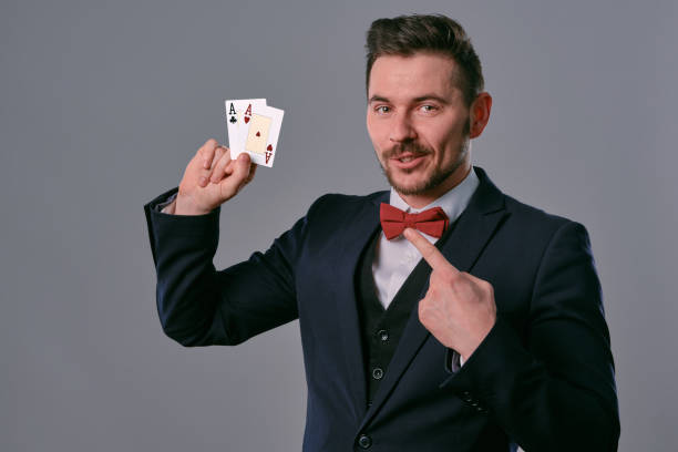 istock Man in black classic suit and red bow-tie showing two playing cards while posing against gray studio background. Gambling, poker, casino. Close-up 1210119816