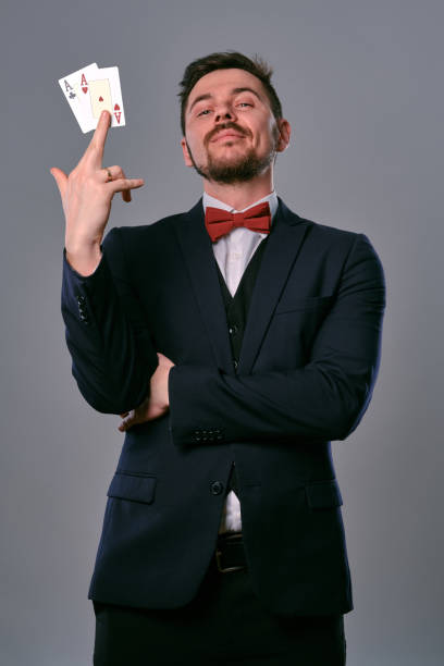 istock Man in black classic suit and red bow-tie showing two playing cards while posing against gray studio background. Gambling, poker, casino. Close-up 1210119832