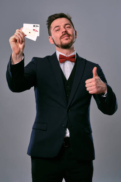 istock Man in black classic suit and red bow-tie showing two playing cards while posing against gray studio background. Gambling, poker, casino. Close-up 1210119847