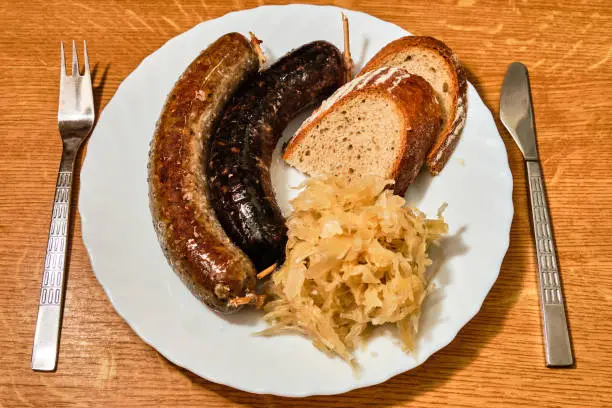 Tasty czech traditional jitrnice, sausage-meat, and jelita, blood sausages, with shredded cabbage and bread on white plate on wooden table with fork and knife top view