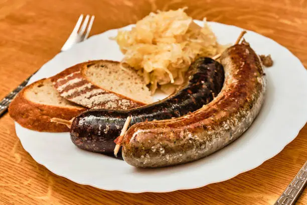 Tasty czech traditional jitrnice, sausage-meat, and jelita, blood sausages, with shredded cabbage and bread on white plate on wooden table with fork and knife
