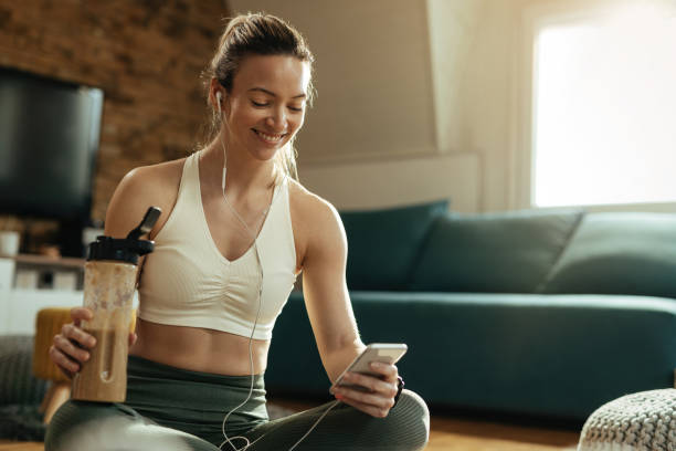 mujer atlética feliz usando el teléfono móvil mientras bebe saqueo en casa. - protein fotografías e imágenes de stock