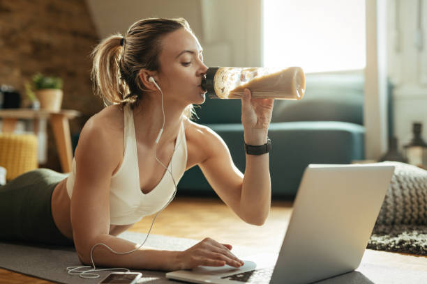 istock Young athletic woman drinking protein shake while using laptop on the floor. 1210121836