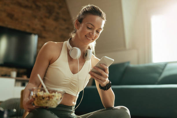 istock Smiling sportswoman reading text message on cell phone while eating salad at home. 1210122044