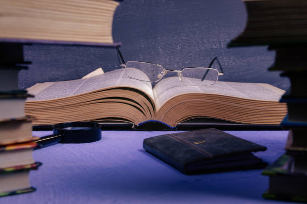 istock Big book with glasses, a magnifier, a notebook on the table through a stack of books with a place for text, writer day, poet, library. 1210123396