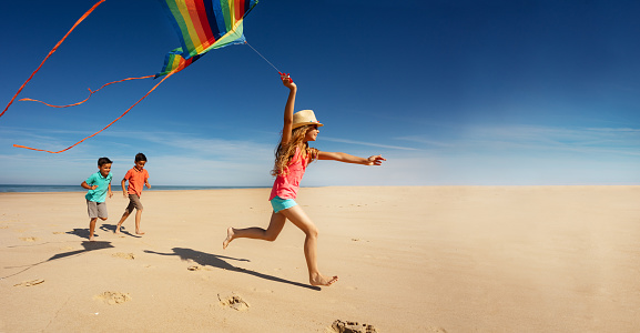 Girl with two boys run on the sand beach holding colorful rainbow stripped kite