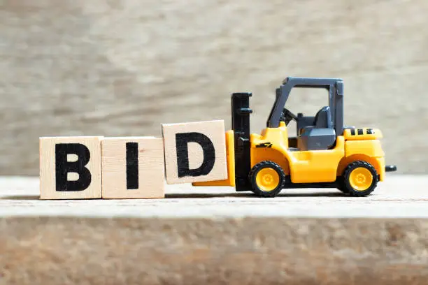 Toy forklift hold letter block d to complete word bid on wood background