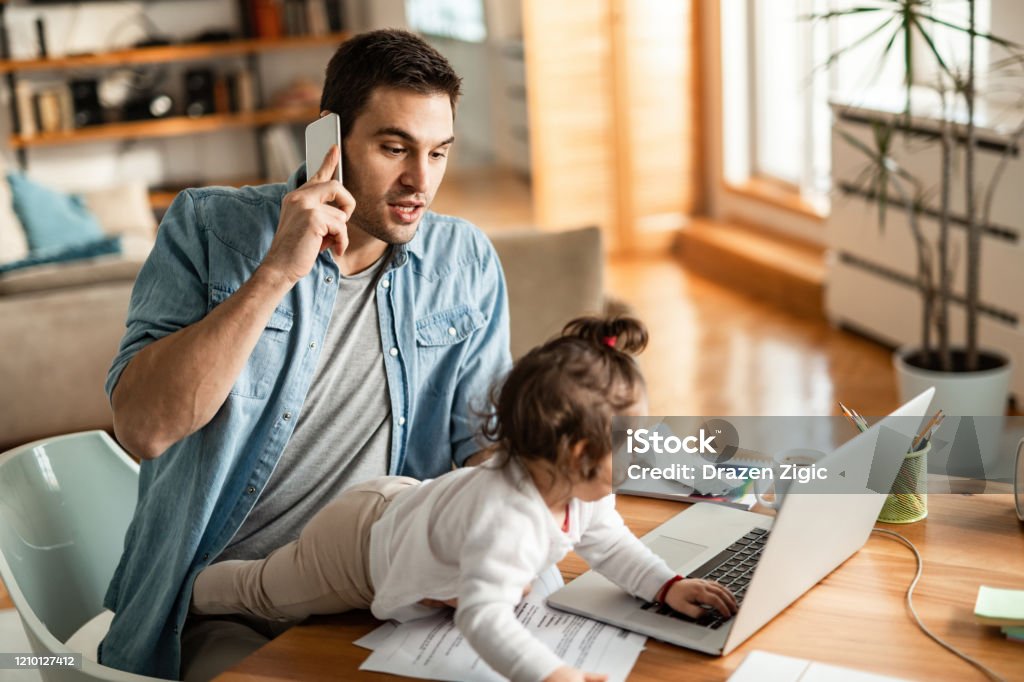 Young working father with small daughter talking on mobile phone at home. Young father babysitting while communicating on cell phone and working at home. Working At Home Stock Photo